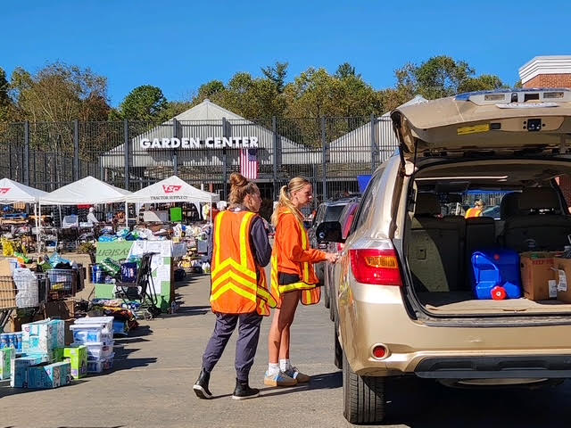 Two volunteers in orange safety vests load supplies into the open back of an SUV at an outdoor distribution site set up in a garden center car park.