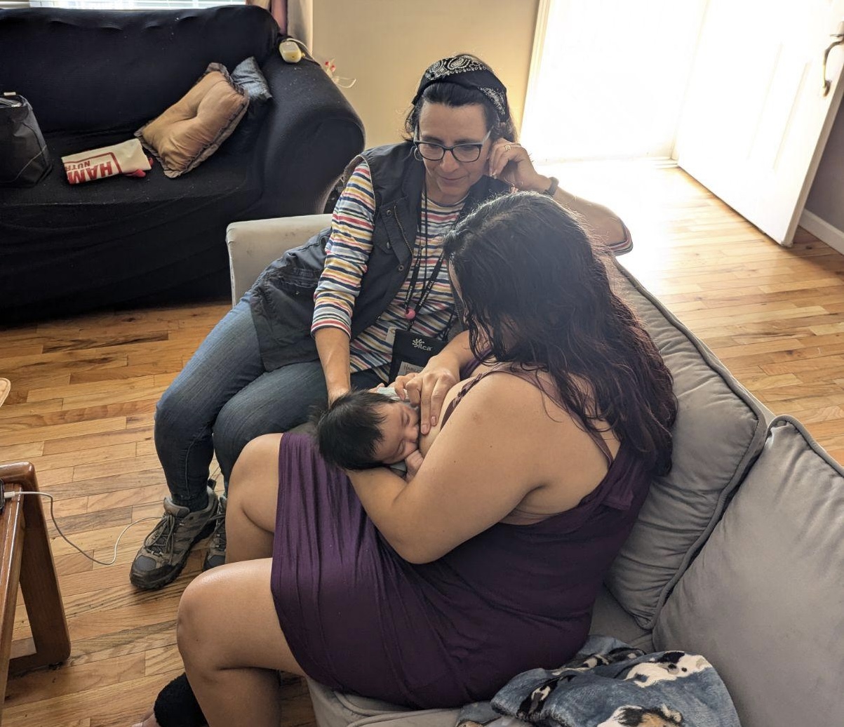 A parent sits on a sofa breastfeeding a baby while a lactation supporter sits beside them, watching and talking.