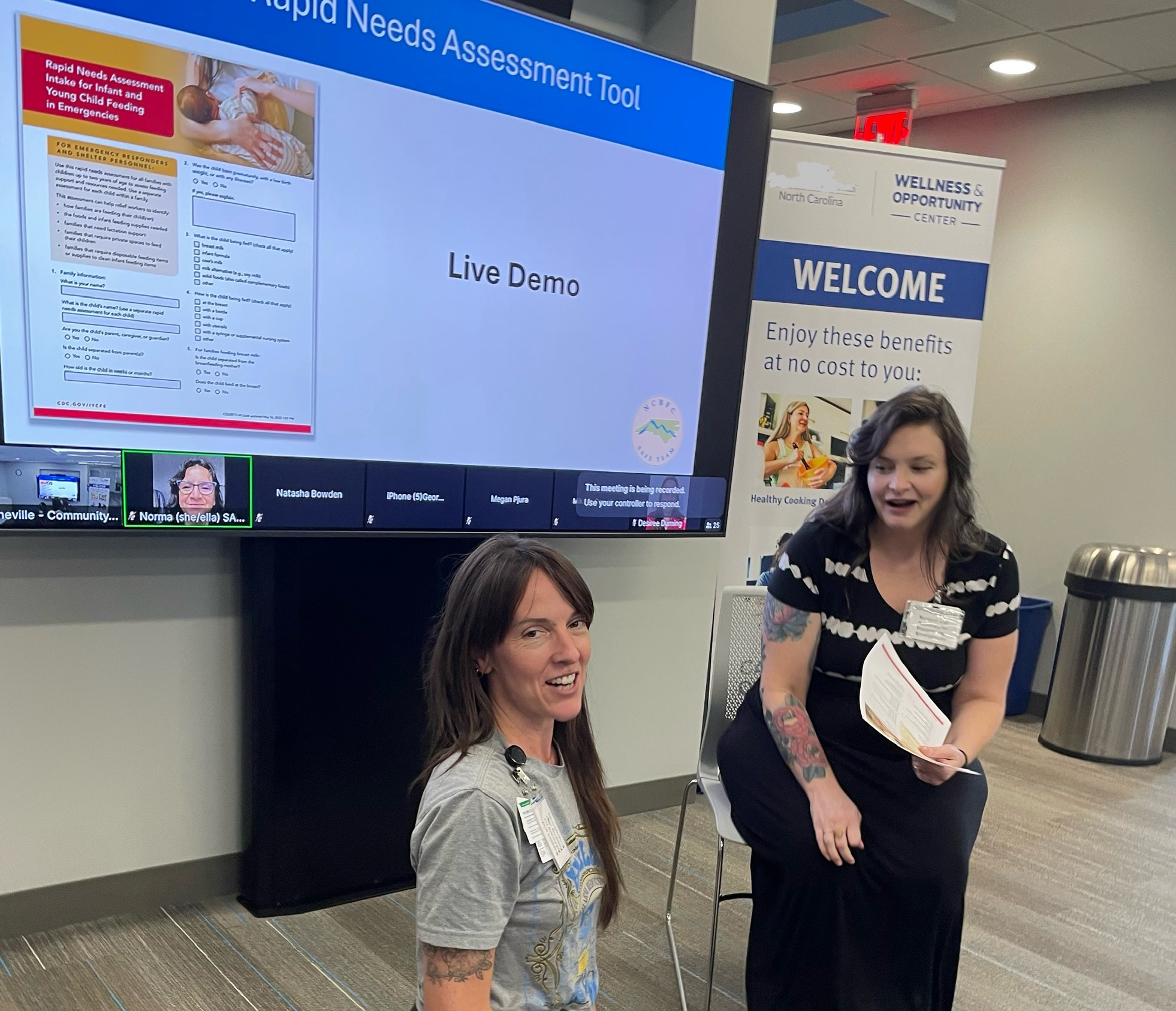 Two SAFE Team trainers stand in front of a projection screen reading Rapid Needs Assessment Tool, Live Demo, during an in-person training in Buncombe County.